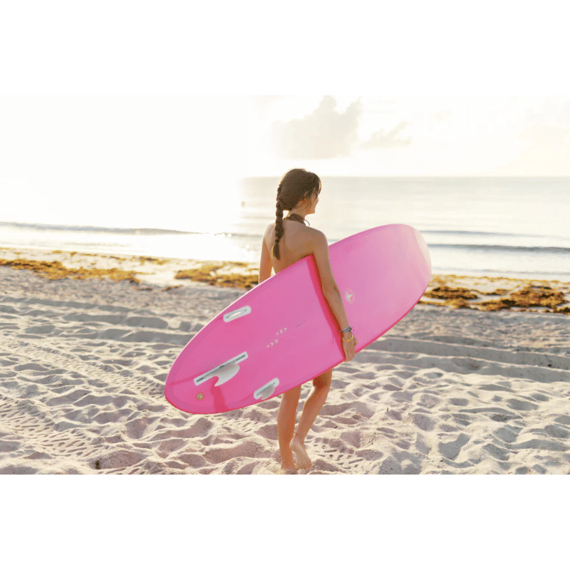 An intermediate surfer carrying a Surfboard Trading Co. ANY DAY Mid Length EPS funboard – Hot Pink