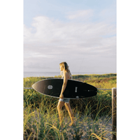 Thumbnail for A person carrying a Surfboard Trading Co. Halligan Carbon Bomb Proof Tech Surfboard | High-Performance Shortboard