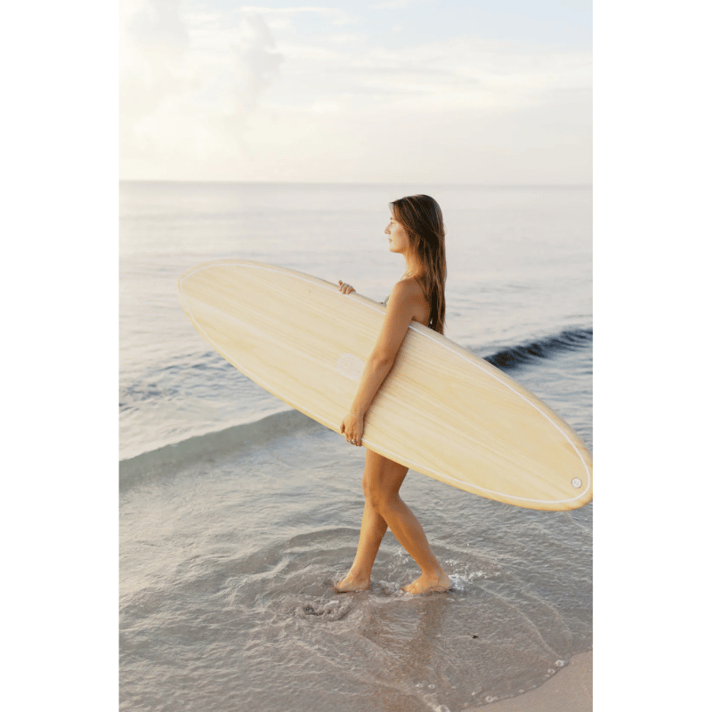 A surfer carrying a Surfboard Trading Co. ANY DAY Mid Length EPS Surfboard – Paulownia Wood