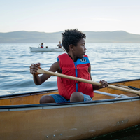 Thumbnail for a child rowing a boat wearing a Mustang Survival Rev Youth Foam Life Jacket red
